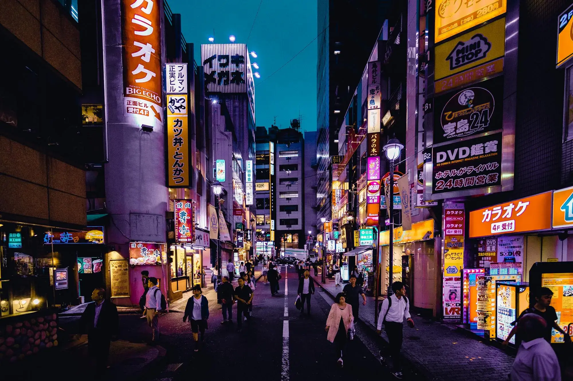 A quiet Tokyo side street at night, neon karaoke signage on both sides.